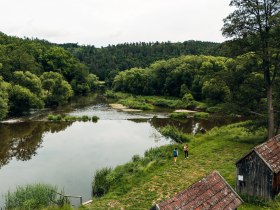 Zwei Personen wandern entlang eines Flussufers, flankiert von dichten B&auml;umen und alten Holzh&uuml;tten, in Drosendorf im Waldviertel.