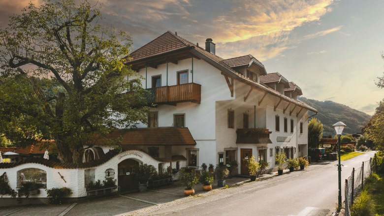 Ein traditionelles Wirtshaus mit weißen Wänden und roten Ziegeldächern, umgeben von Bäumen und einer ruhigen Straße.