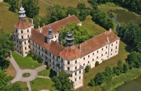 Luftaufnahme eines historischen Schlosses mit zwei T&uuml;rmen und einem Innenhof, umgeben von B&auml;umen und einem Teich.