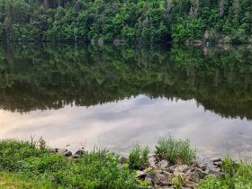 Ein ruhiger See mit bewaldeten H&uuml;geln im Hintergrund, die sich im Wasser spiegeln.