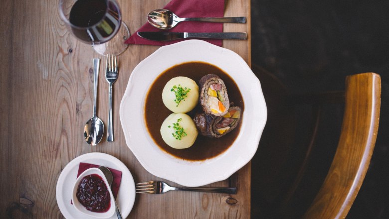 A plate with beef roulade and two potato dumplings in sauce, next to it a glass of red wine and cranberries on a wooden table.