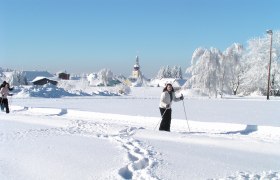 Zwei Personen beim Langlaufen in einer verschneiten Landschaft mit einer Kirche im Hintergrund.