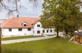 Ein weißes Gebäude mit rotem Ziegeldach, umgeben von Bäumen und einer Wiese, mit einem Spielplatz im Vordergrund.