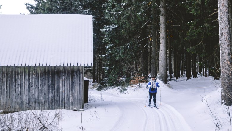 Person beim Skilanglauf im verschneiten Wald neben einer Holzh&uuml;tte.