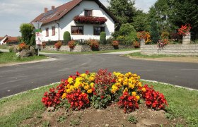 Ein weißes Haus mit roten Dachziegeln und Blumenschmuck, umgeben von bunten Blumenbeeten und grüner Landschaft.