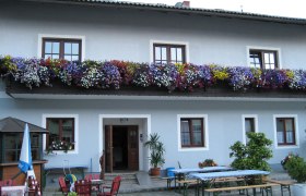 Gasthof mit blauer Fassade, Blumenkästen am Balkon und Biergartenmöbeln im Vordergrund.