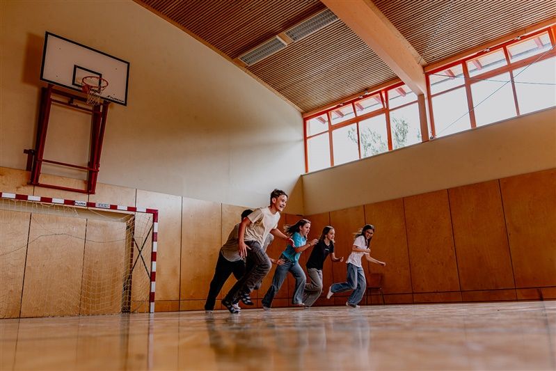 Fünf Jugendliche rennen in einer Turnhalle mit Basketballkorb und Handballtor.