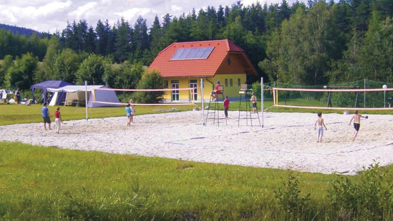 Menschen spielen Volleyball auf einem Sandplatz vor einem gelben Gebäude mit rotem Dach, umgeben von Bäumen.
