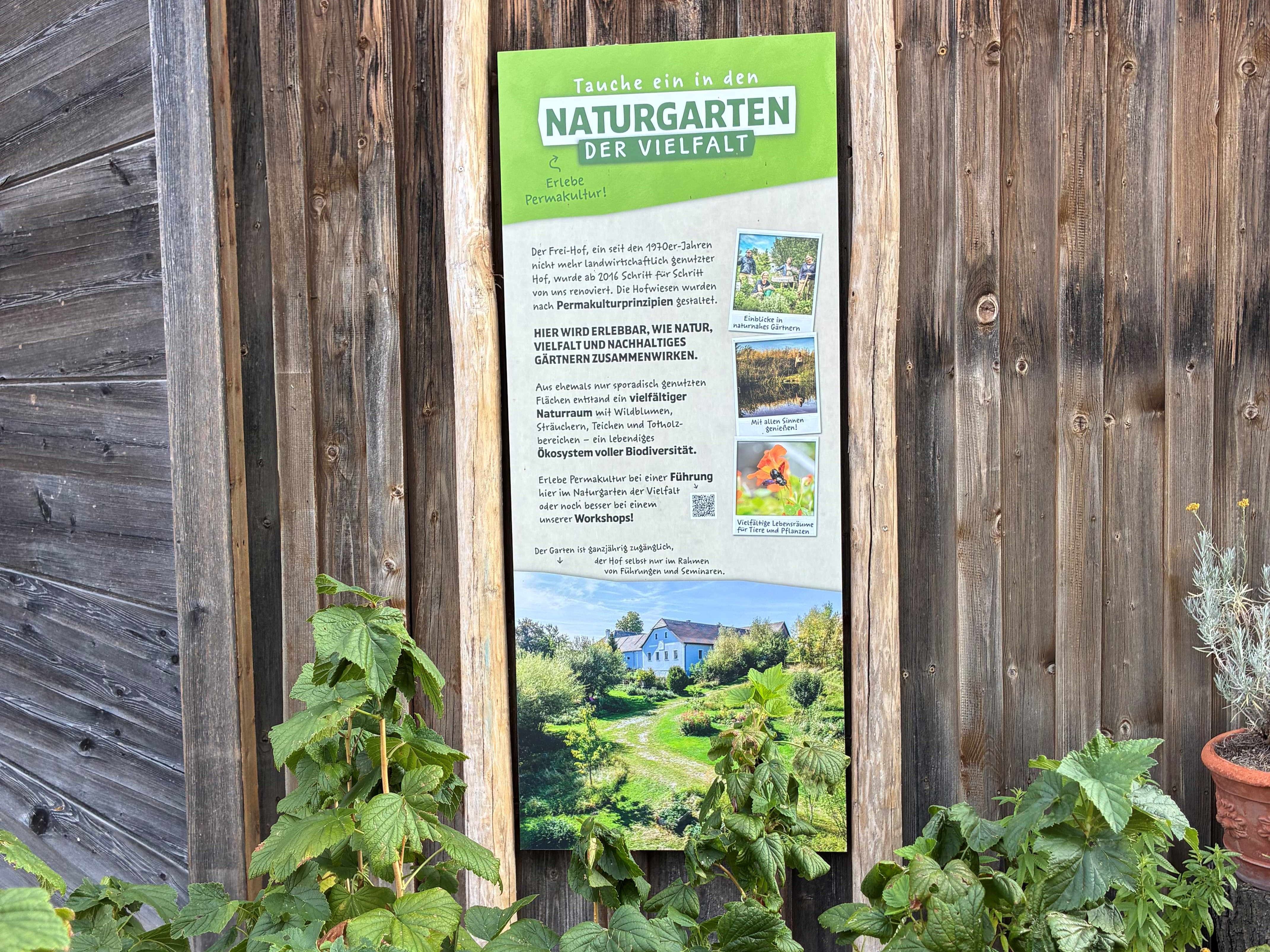 Ein Schild mit der Aufschrift 'Naturgarten der Vielfalt' an einer Holzwand, umgeben von Pflanzen.