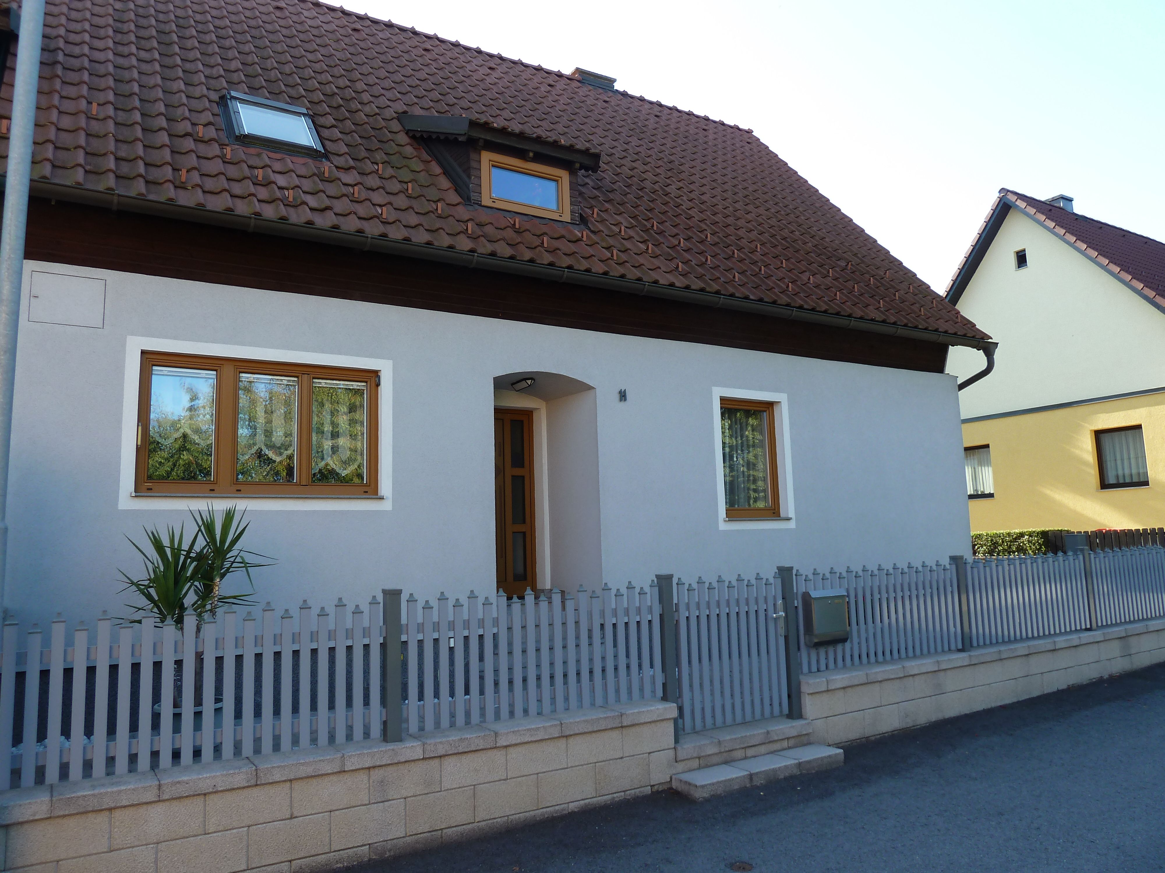 Ein Einfamilienhaus mit grauer Fassade, braunem Dach und Holztüren. Ein kleiner Zaun umgibt das Grundstück.