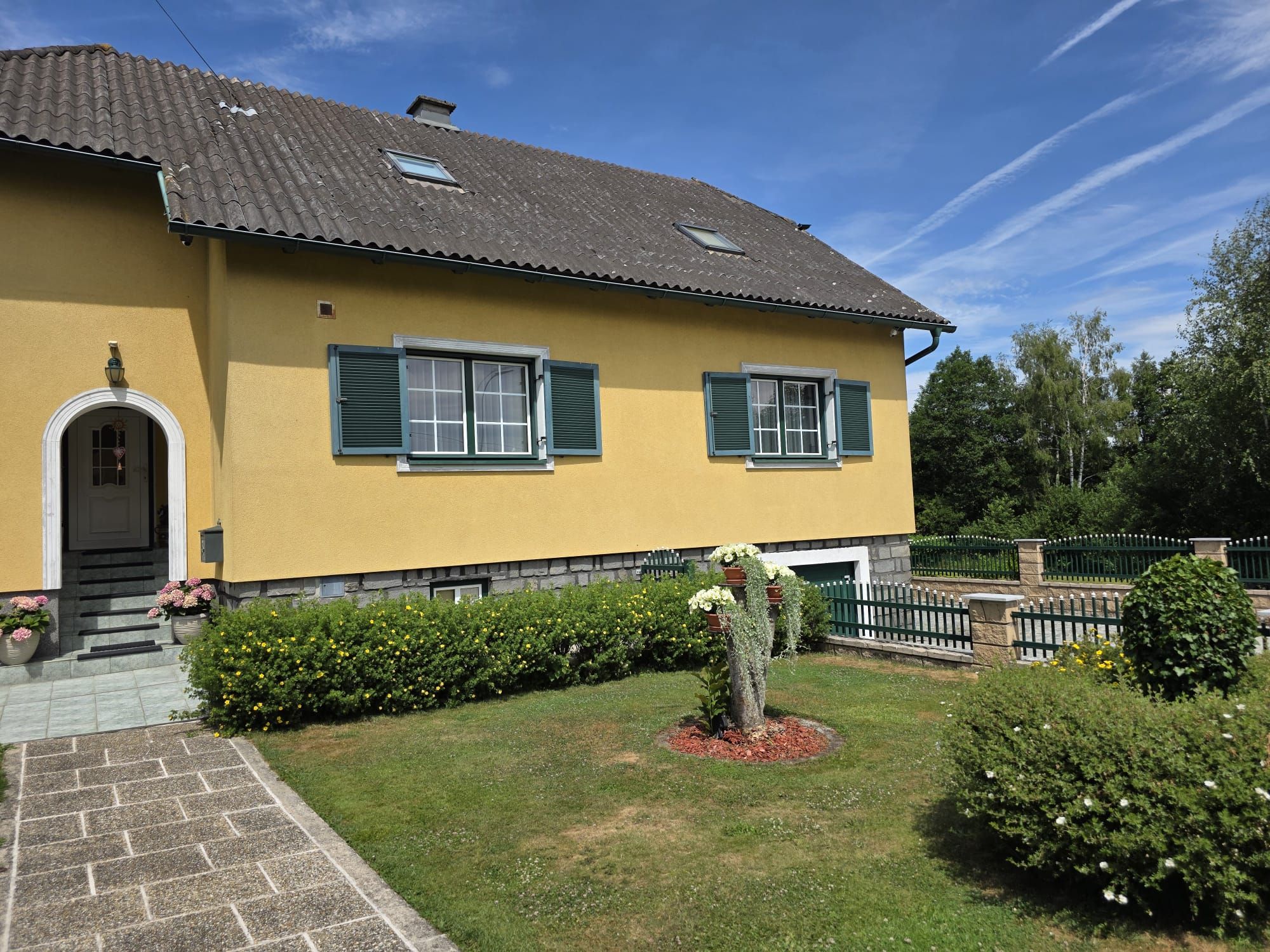 Gelbes Einfamilienhaus mit grünem Fensterladen und gepflegtem Garten bei sonnigem Wetter.