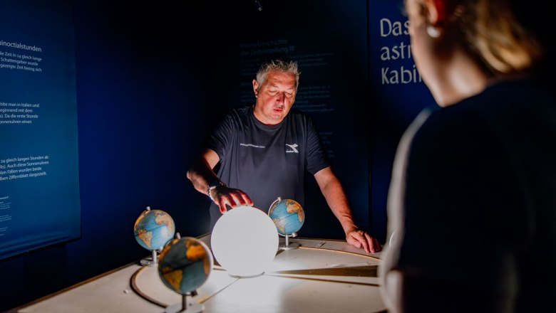 A man explains a model with globes and a glowing sphere in a dark room.