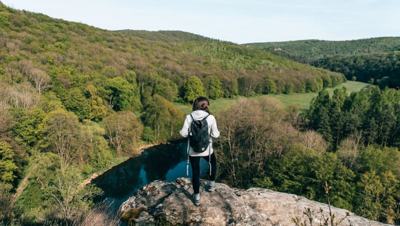 Person mit Rucksack steht auf einem Felsen und blickt auf bewaldete Landschaft im Nationalpark Thayatal.