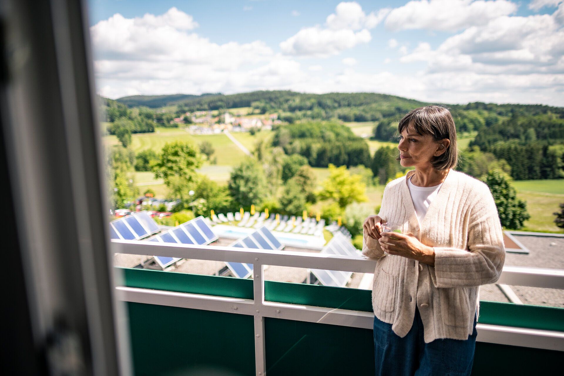 Person mit einer Tasse auf dem Balkon des Gesundheits- und Rehabilitationszentrums Moorheilbad Harbach im Waldviertel mit Blick auf die grüne Hügellandschaft.
