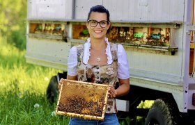 Eine Frau in traditioneller Kleidung hält eine Bienenwabe vor einem Bienenwagen.