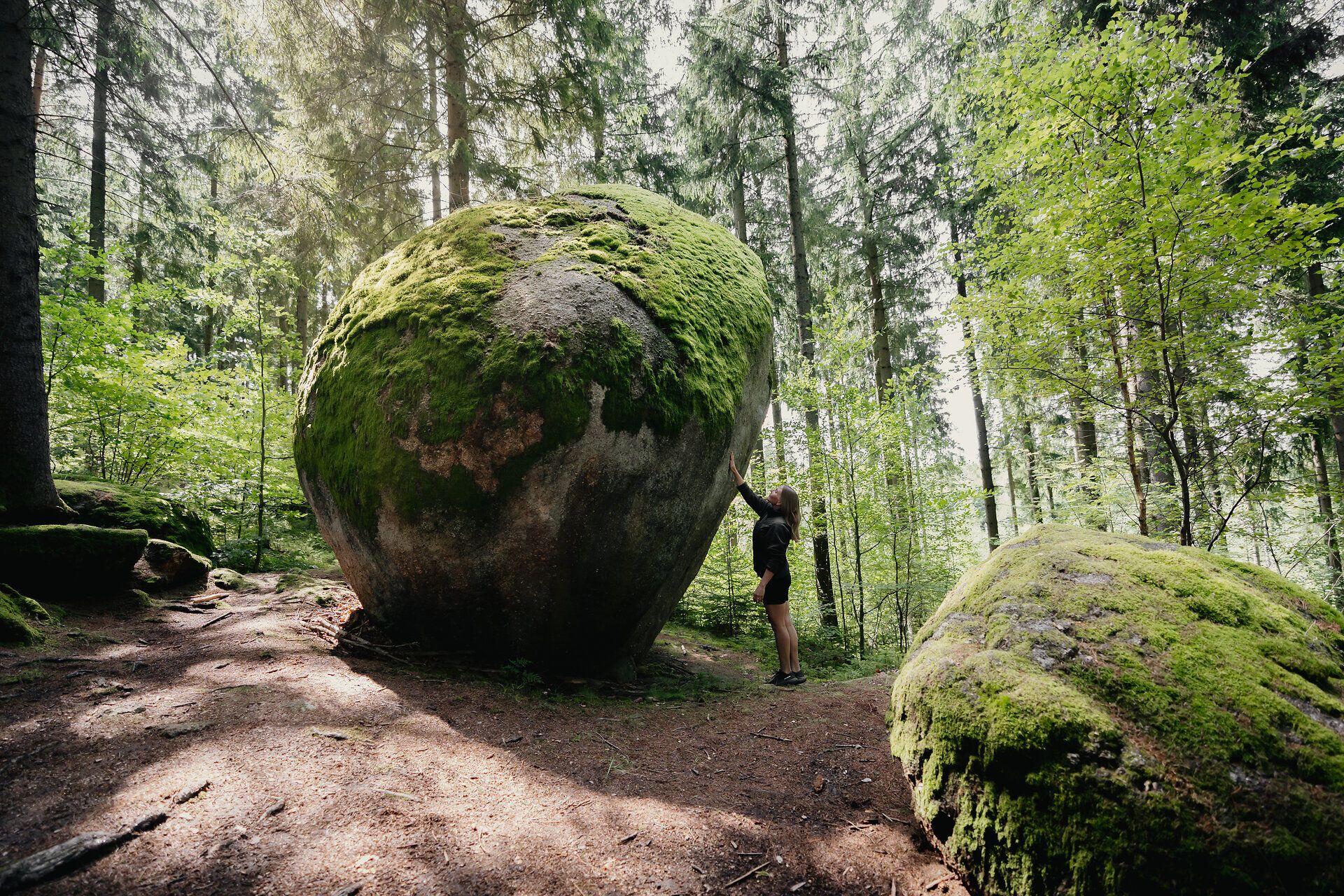 Person steht im Wald vor einem großen, kugelförmigen Felsen, der von Moos bedeckt ist; die sogenannte Weltkugel liegt in der Kraftarena Groß Gerungs, umgeben von hohen Bäumen.