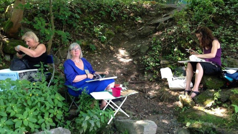 Drei Frauen sitzen im Freien und zeichnen in einem bewaldeten Gebiet.