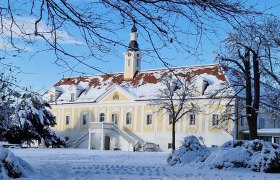 Schloss Haindorf im Winter mit Schnee bedeckt, blauer Himmel und kahle Bäume im Vordergrund.