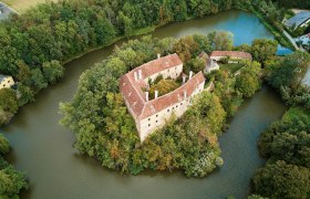 Luftaufnahme einer von Wasser umgebenen Burg mit roten D&auml;chern und umgebenden B&auml;umen.