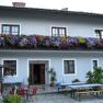 Gasthof mit blauer Fassade, Blumenkästen am Balkon und Biergartenmöbeln im Vordergrund.