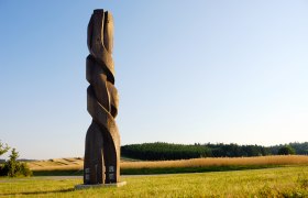 Holzskulptur auf einem Feld mit blauem Himmel im Hintergrund.