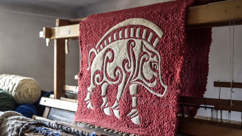 Ein roter Wandteppich mit einem weißen, stilisierten Tiermotiv auf einem Webstuhl.