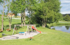 Spielplatz mit Schaukel, Rutsche und Wippe neben einem Teich in einer grünen Landschaft.
