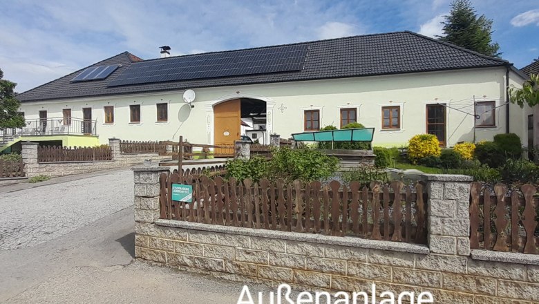Außenanlage – Ferienwohnung rechts, © Erika Polt Ein großes Gebäude mit gelber Fassade und schwarzem Dach, umgeben von einem Garten und einem Holzzaun. Der Himmel ist blau mit einigen Wolken.