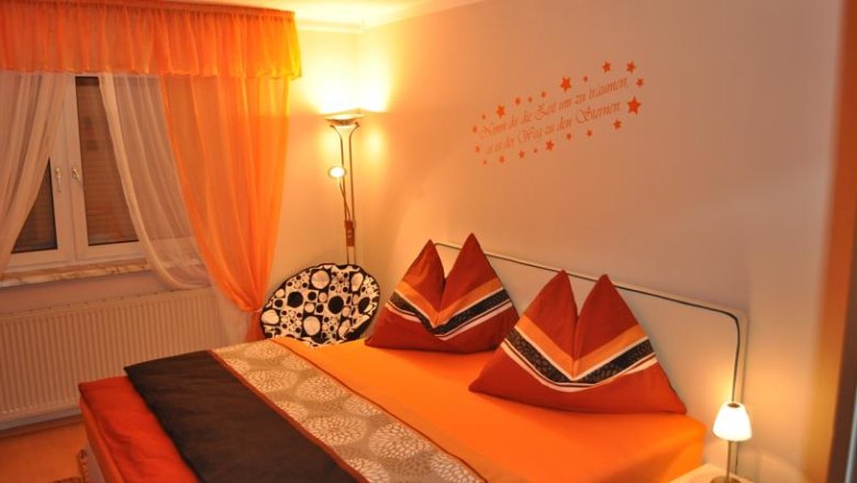 Gemütliches Schlafzimmer mit orangefarbener Bettwäsche und Vorhängen, dekorativen Kissen und Wandtattoo.