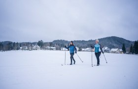 Zwei Personen beim Langlaufen auf einer schneebedeckten Fl&auml;che vor einem Dorf und bewaldeten H&uuml;geln.
