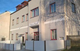 Außenansicht der Pension Brandl mit grauer Fassade und roten Fensterrahmen.