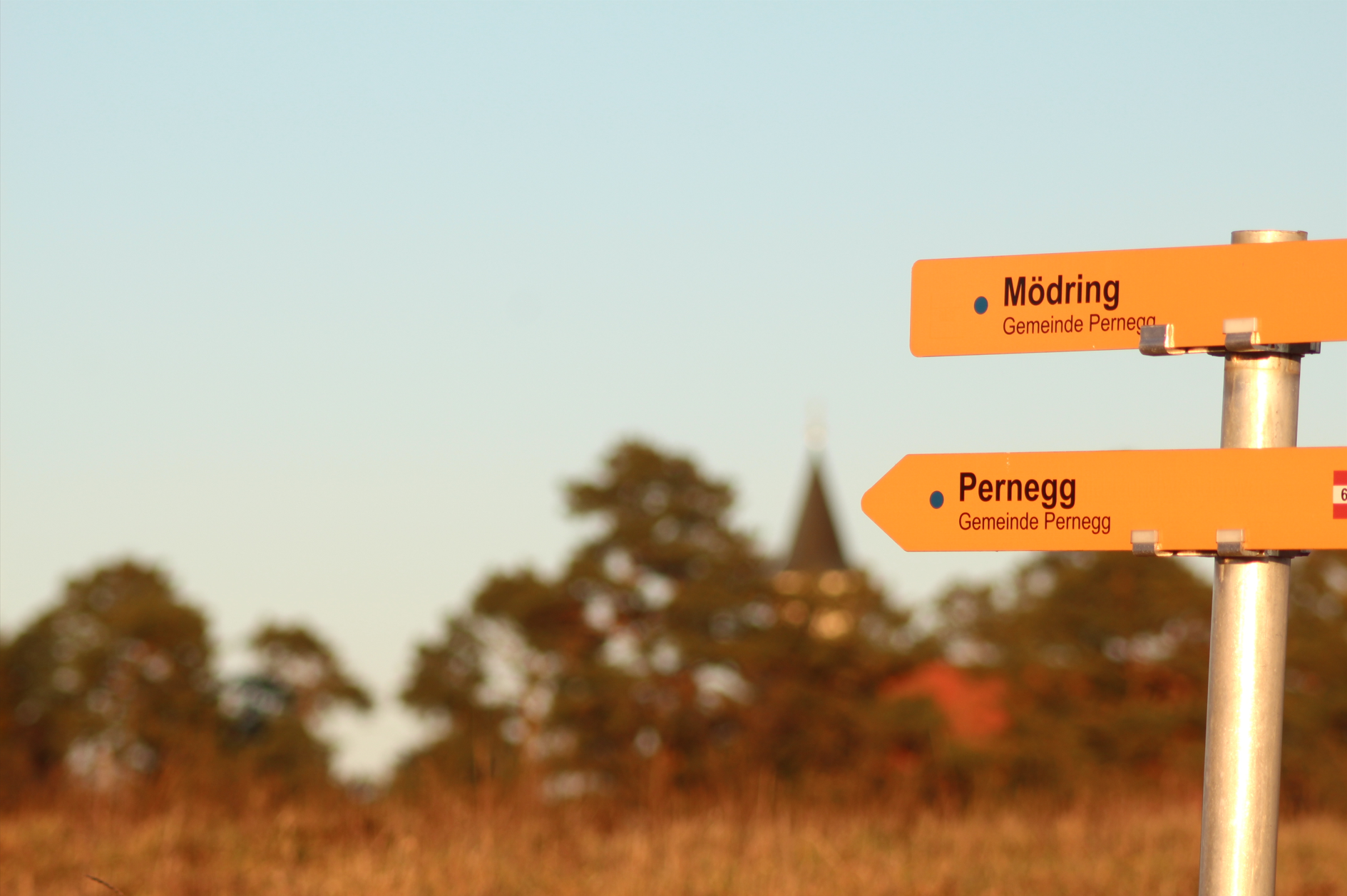 Wegweiser mit den Zielen Mödring und Pernegg vor unscharfem Hintergrund mit Bäumen und Kirchturm.