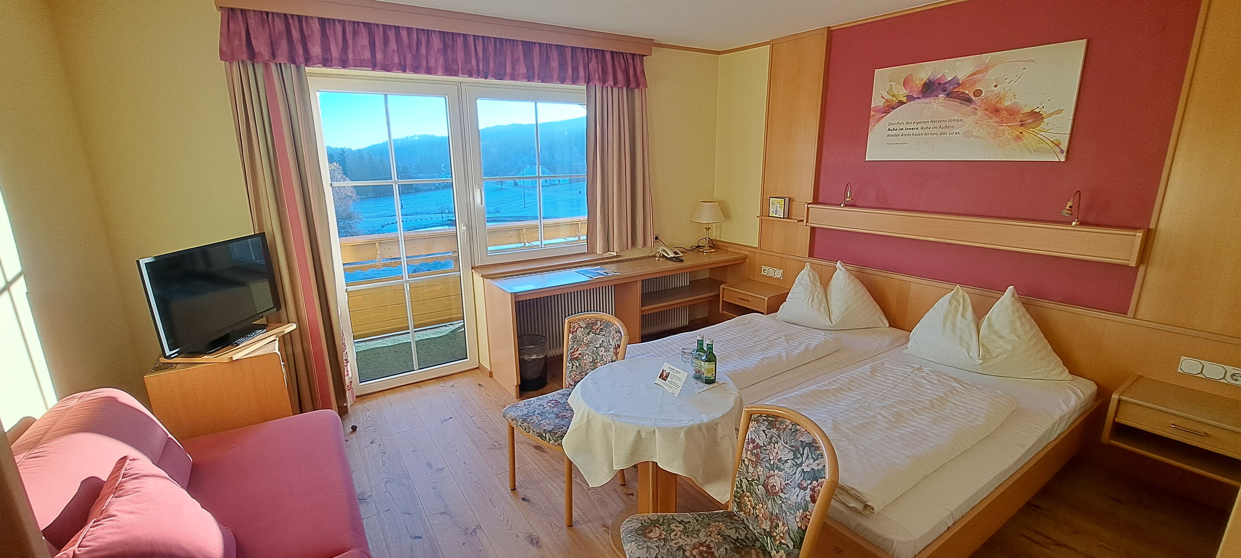 Gemütliches Hotelzimmer mit Doppelbett, Tisch, Stühlen und Fernseher. Fenster mit Blick auf Berge.