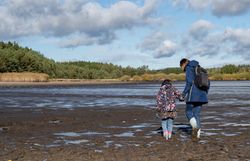 Ein strahlend blauer Himmel spiegelt sich im ruhigen Wasser des Bruneiteichs, während ein Erwachsener und ein Kind Hand in Hand über den schimmernden, schlammigen Boden spazieren. Die sanfte Brise und die umgebende Natur schaffen eine friedliche Atmosphäre, die zum Verweilen einlädt.