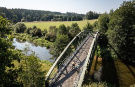 Die sanfte Br&uuml;cke schwingt &uuml;ber den glitzernden Fluss, umgeben von &uuml;ppigem Gr&uuml;n und der Ruhe der Natur. Radfahrer genie&szlig;en die frische Luft und die malerische Landschaft, w&auml;hrend die Sonne sanft auf die Wiesen scheint.