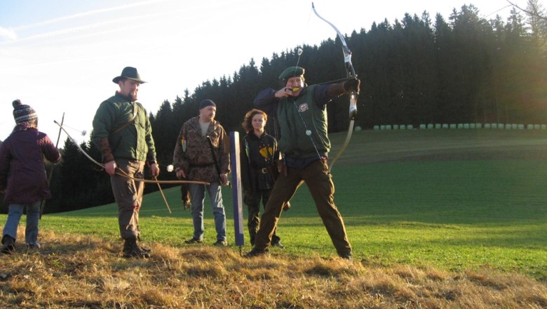 Gruppe von Menschen beim Bogenschie&szlig;en auf einer Wiese vor einem Wald.