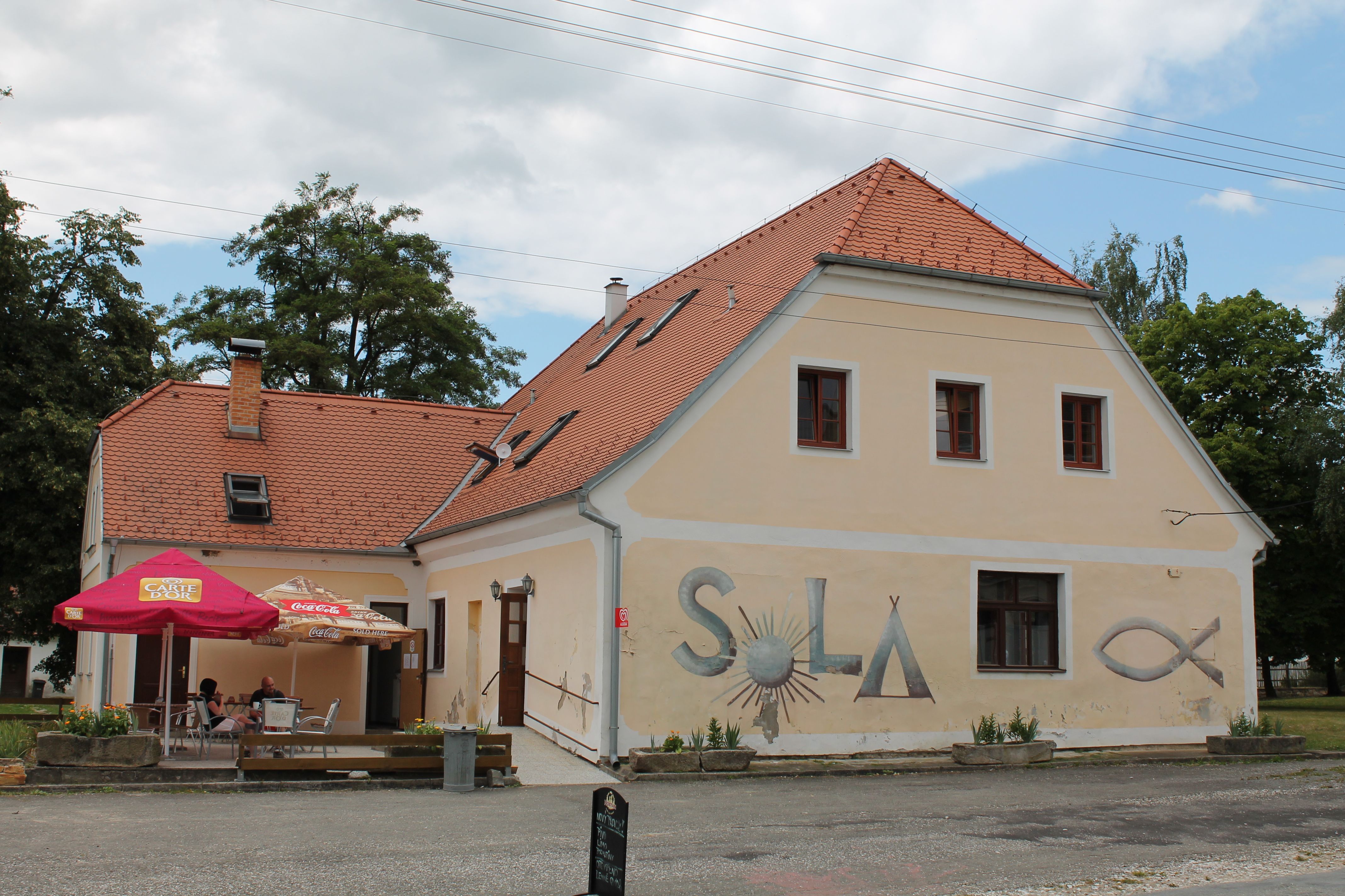 Ein gelbes Gebäude mit rotem Ziegeldach und der Aufschrift 'SOLA' an der Wand. Vor dem Gebäude stehen Sonnenschirme und Tische.