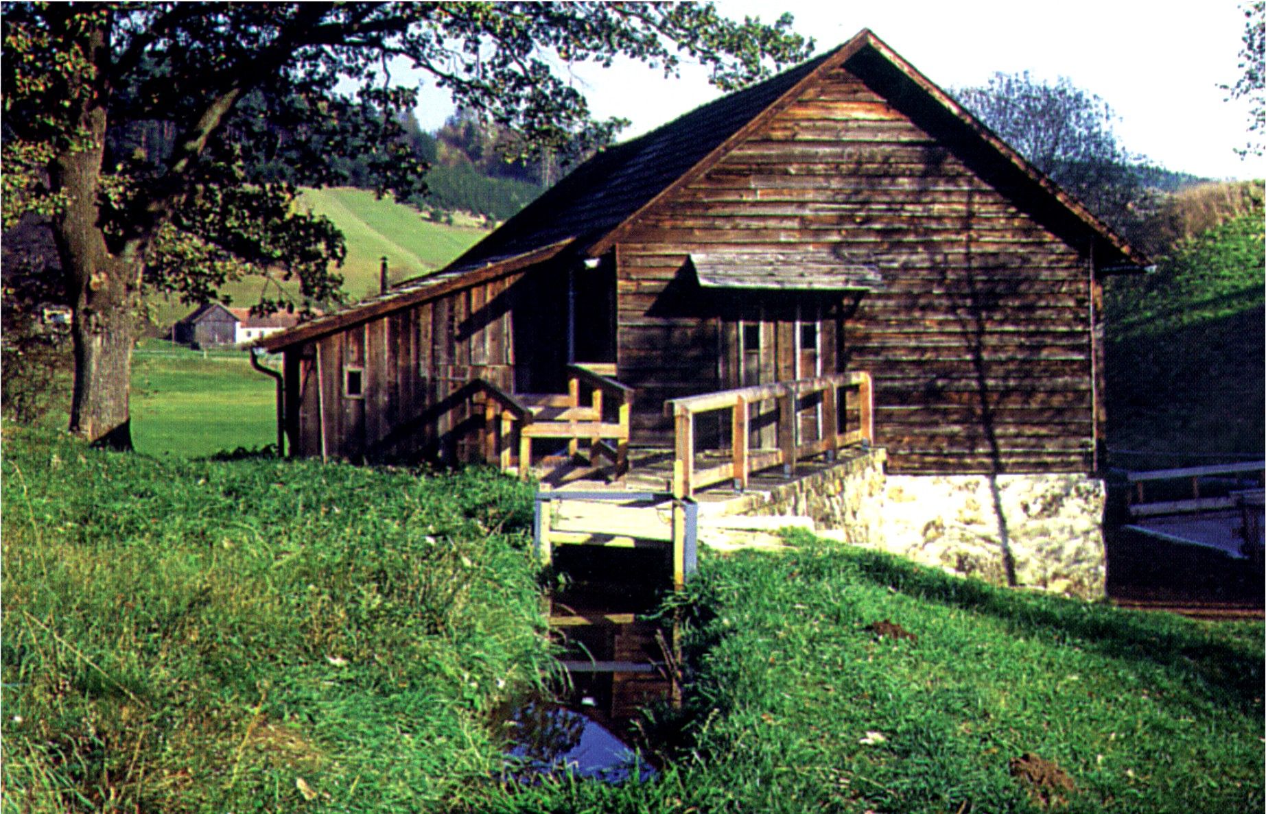 Altes Holzgebäude mit Brücke und Bach in ländlicher Umgebung.