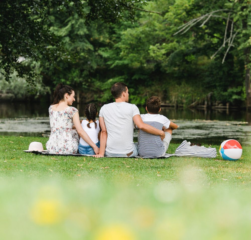 Eine Familie genießt einen entspannten Tag am Ufer eines ruhigen Gewässers, umgeben von üppigem Grün und blühenden Wiesen. Die Kinder spielen mit einem bunten Ball, während die Erwachsenen die friedliche Atmosphäre und die malerische Landschaft in vollen Zügen auskosten.