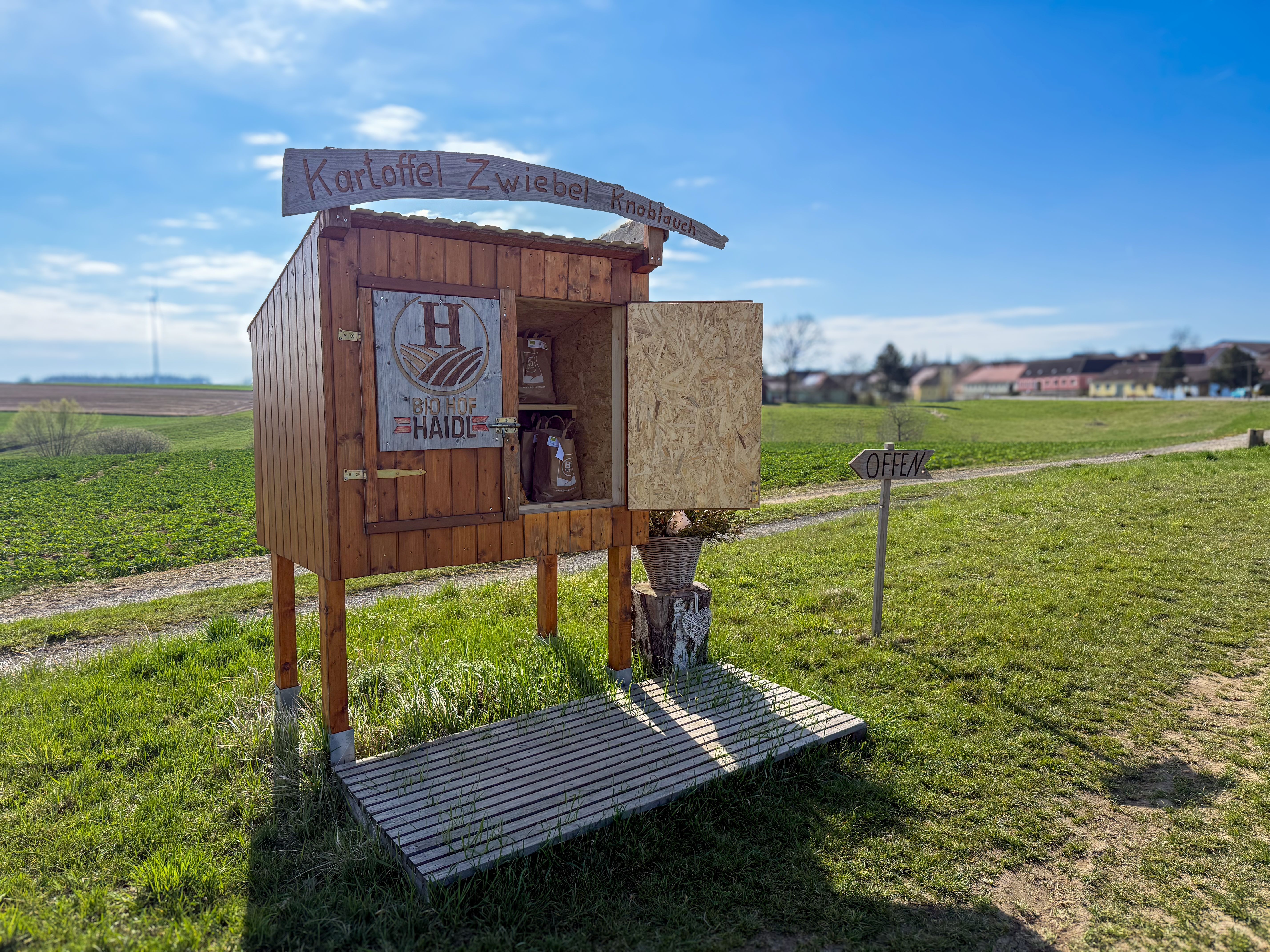 Ein kleiner Verkaufsstand auf einem Feldweg mit der Aufschrift 'Kartoffel Zwiebel Knoblauch' und 'Bio Hof Haide'. Ein Schild zeigt 'Offen'.