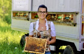 Eine Frau in traditioneller Kleidung hält eine Bienenwabe vor einem Bienenwagen.