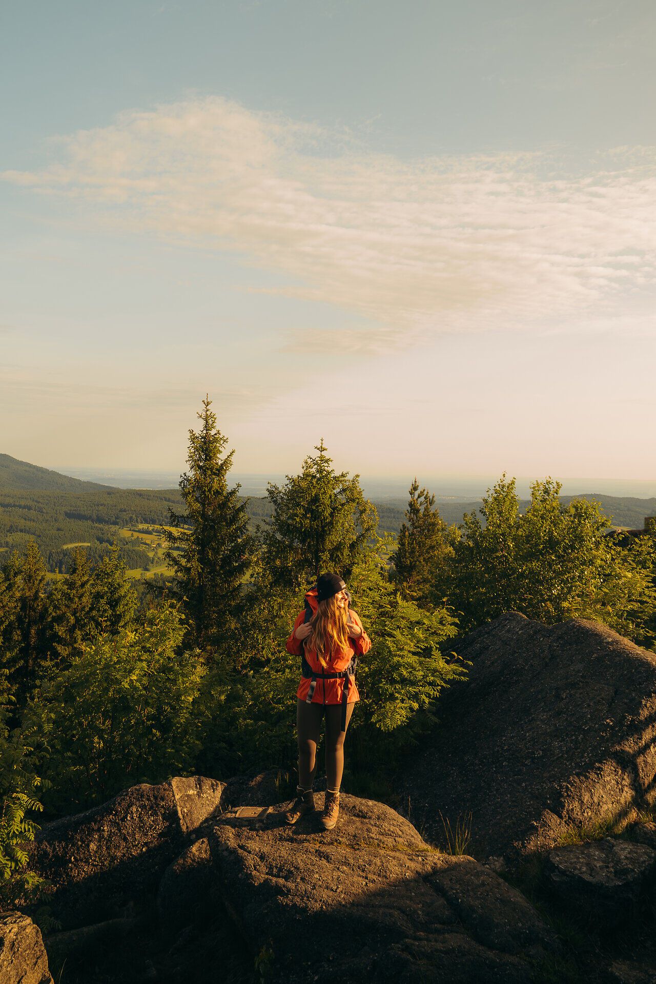 Ein Wanderer steht auf einem Felsen mit Ausblick auf bewaldete Hügel, aufgenommen im Waldviertel, gekrönt von einem klaren Himmel und dem Licht der aufgehenden Sonne.
