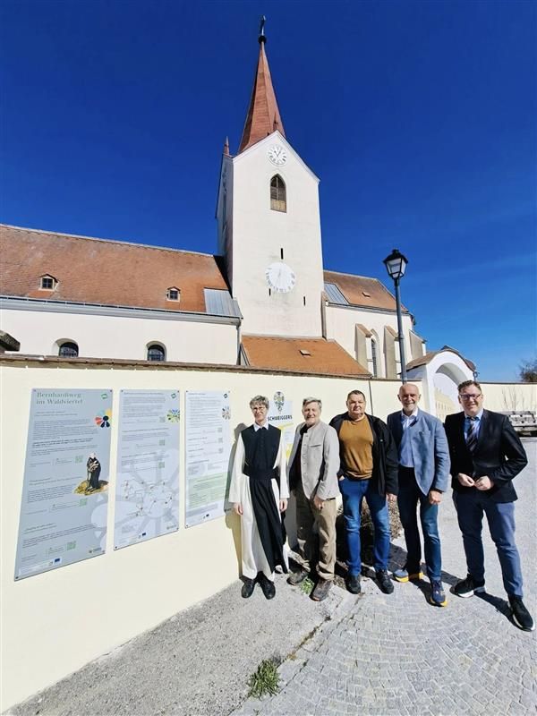 Fünf Männer neben den neuen Infotafeln des Bernhardiwegs. Dahinter eine Kirche in gelb. 