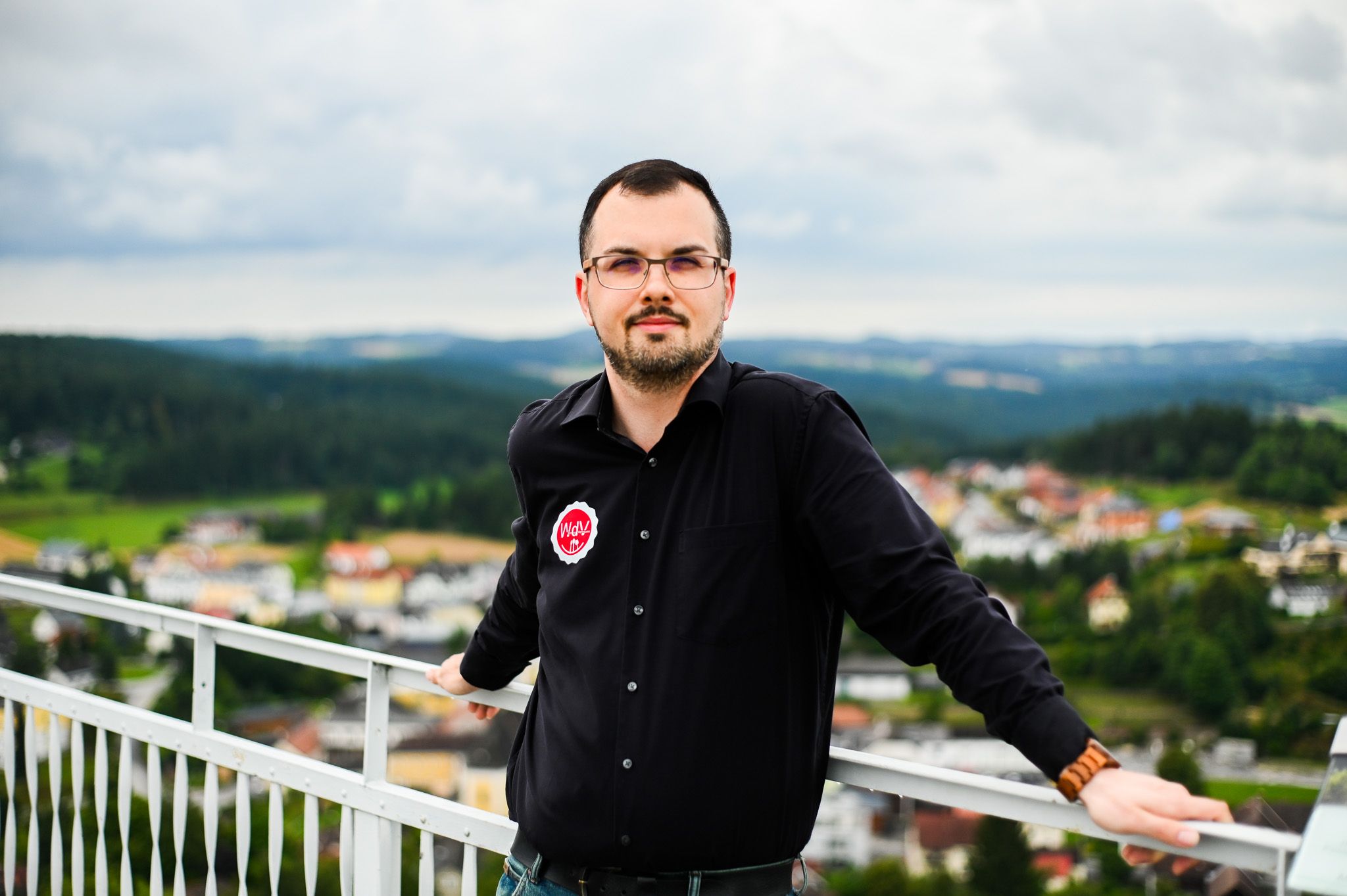 Ein Mann mit Brille und schwarzem Hemd steht an einem Geländer mit einer Landschaft im Hintergrund.
