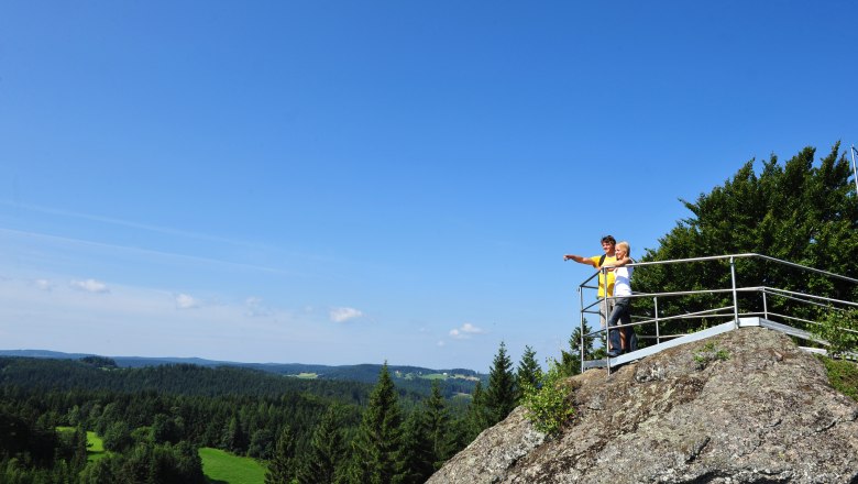 Zwei Personen stehen auf einem Aussichtspunkt auf einem Felsen und blicken in die Ferne über eine bewaldete Landschaft.