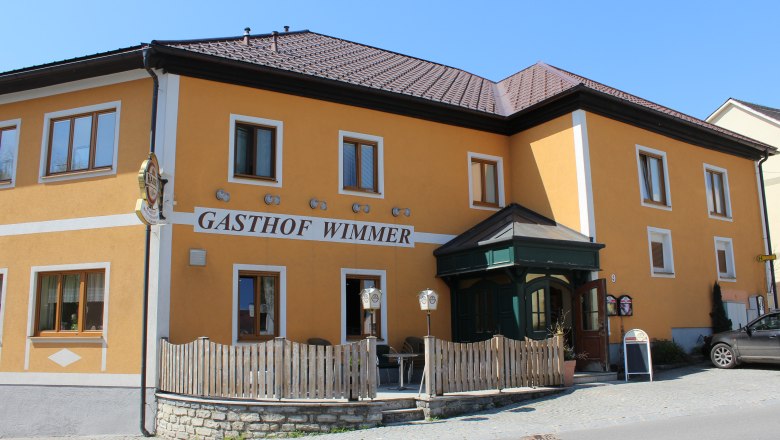 Gelbes Gebäude mit der Aufschrift 'Gasthof Wimmer', Holzzaun und Eingangstür.