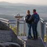 A couple stands on a viewing platform with a view of a vast landscape.