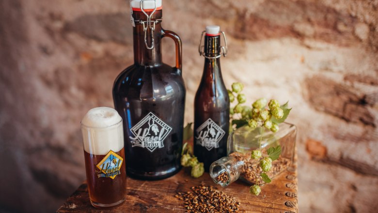 Bierflaschen und Glas mit Haselbräu-Logo, Hopfen und Malz auf Holztisch.
