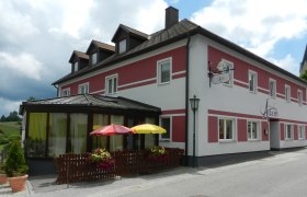 Ein Gasthaus mit roter und weißer Fassade, Terrasse mit Sonnenschirmen und Blumendekoration.