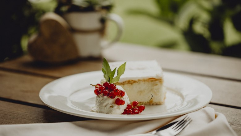 Ein Teller mit einer Cremeschnitte, Sahne, roten Johannisbeeren und einem Minzblatt auf einem Holztisch.